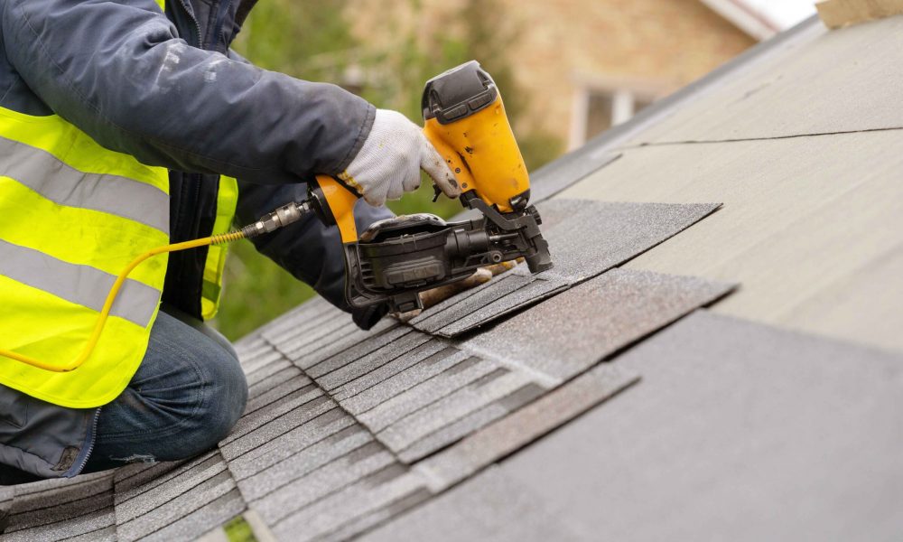 Roofer worker in special protective work wear and gloves, using air or pneumatic nail gun and installing asphalt or bitumen shingle on top of the new roof under construction residential building